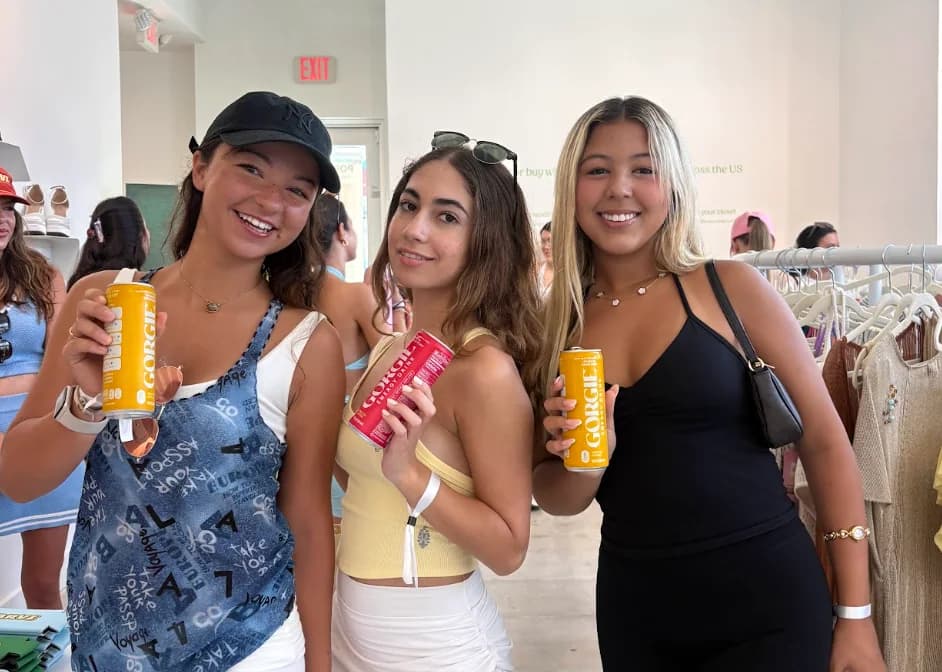 Three women smiling together and holding canned drinks at a shop event.