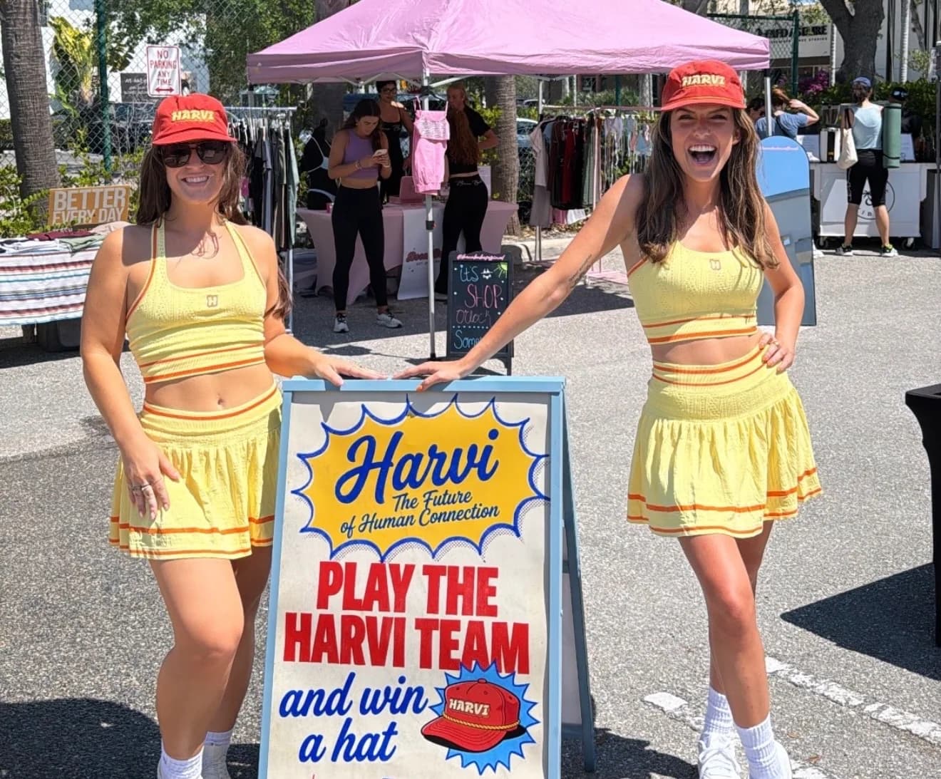 Two women in matching yellow Harvi outfits standing beside a Harvi team sign at an outdoor market.